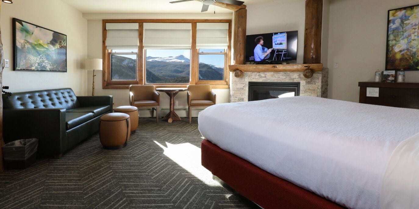 A hotel room with a large bed, a sofa, two chairs, a fireplace, a TV, and a view of mountains through large windows.