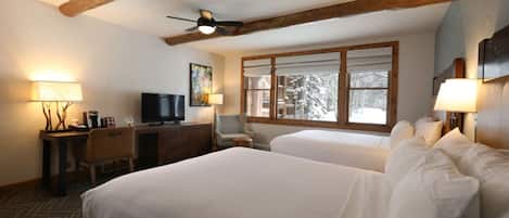 A hotel room with two beds, a desk with a television, and a window with a view of snow-covered trees.