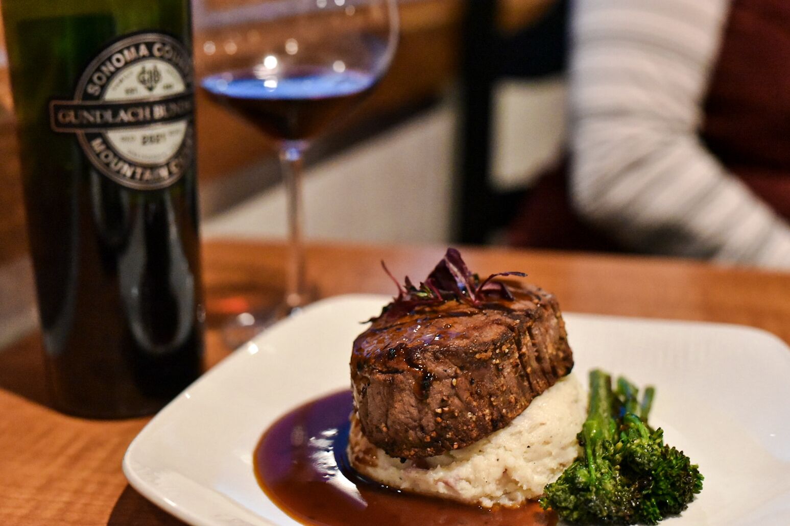 Ein Teller mit Essen, darunter ein Steak, Kartoffelpüree und Brokkoli, dazu eine Weinflasche und ein Glas Rotwein.