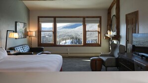 Une chambre d’hôtel avec un grand lit, un bureau, une chaise, une télévision et une vue sur des montagnes enneigées, visible à travers de grandes fenêtres.