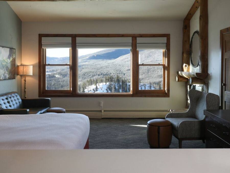 Habitación de hotel con una cama grande, un escritorio, una silla, un televisor y vistas a montañas nevadas a través de amplias ventanas.