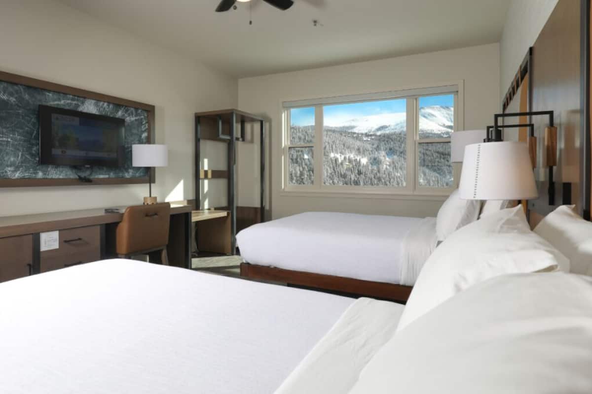 Een hotelkamer met een bed, een bureau, een stoel, een televisie, een raam met uitzicht op een besneeuwde berg en een lamp.