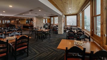 Un restaurant espaiós amb diverses taules i caires per dinar, una zona de bar i grans finestres que ofereixen vista a un paisatge nevós.
