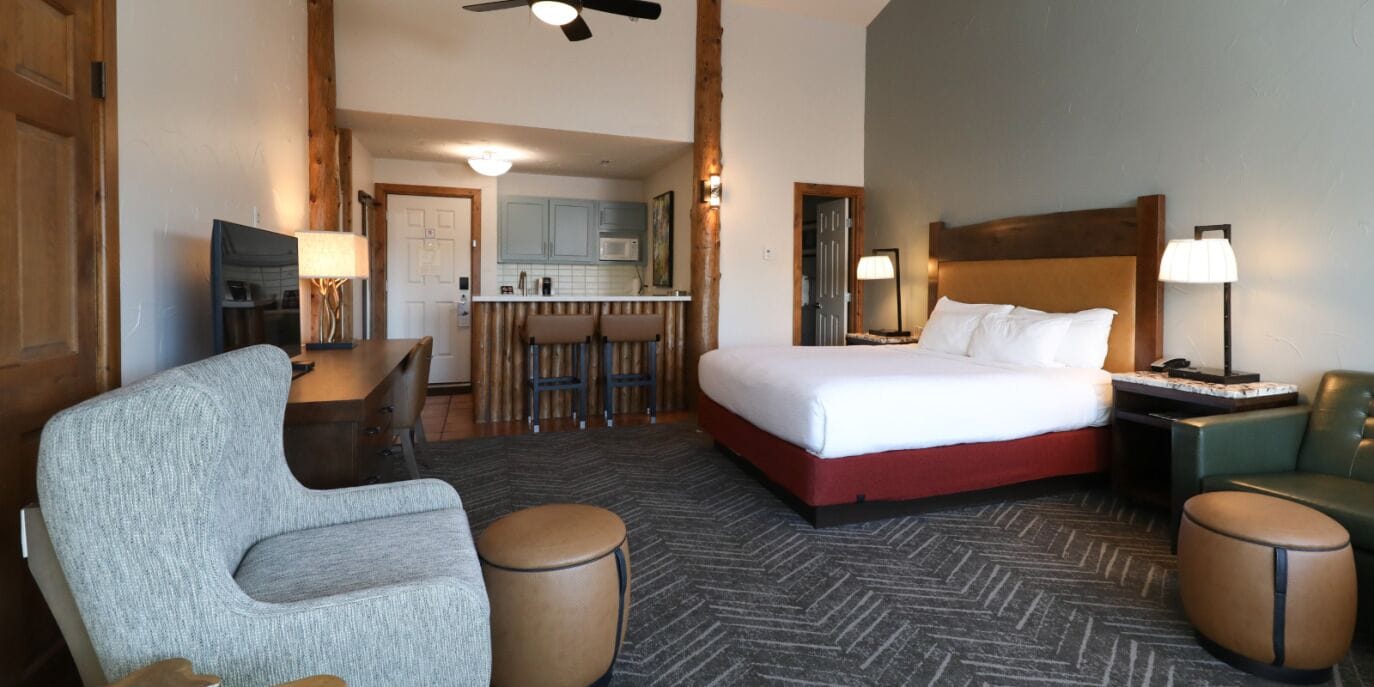 A hotel room with a bed, a desk, a chair, and a kitchenette in the background.
