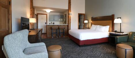 A hotel room with a bed, a desk, a chair, and a kitchenette in the background.