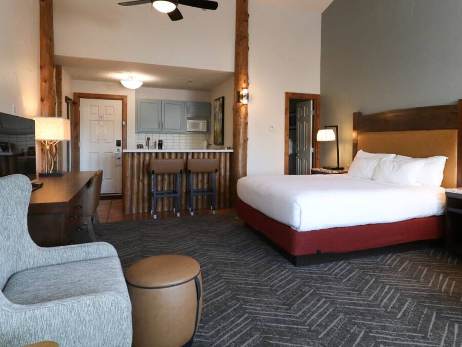 A hotel room with a bed, a desk, a chair, and a kitchenette in the background.