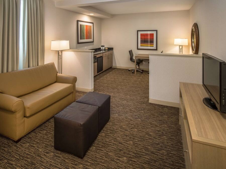 A hotel room with a sofa, ottoman, desk, television, and artwork on the walls.