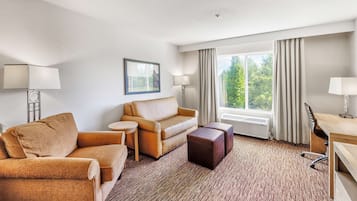Habitación de hotel con sofá color beige, una mesita redonda pequeña, un reposapiés oscuro, un escritorio con silla y un ventanal con cortinas.