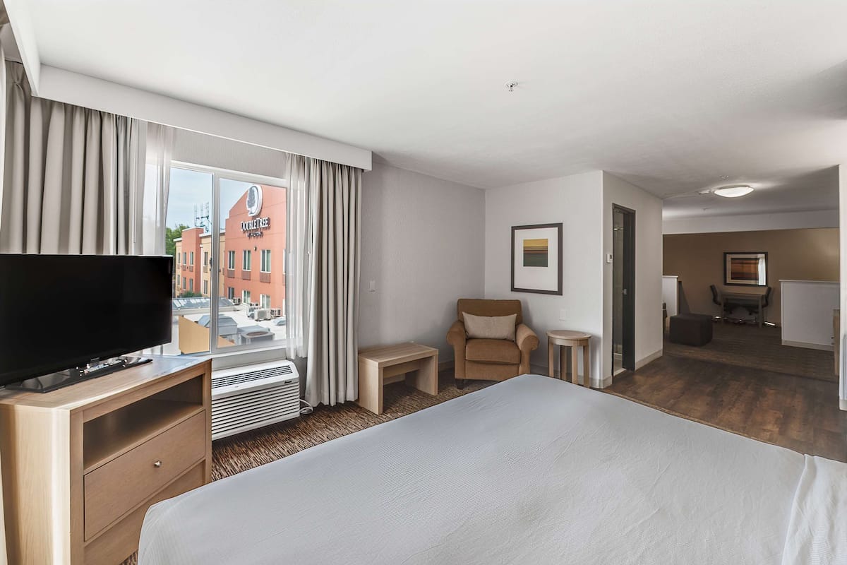 Una habitación de hotel con cama, televisión, silla, mesita y ventana con cortinas.