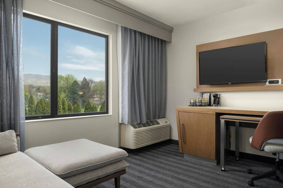 Een hotelkamer met een bed, een bureau, een stoel, een televisie en een raam met uitzicht op bomen en heuvels.