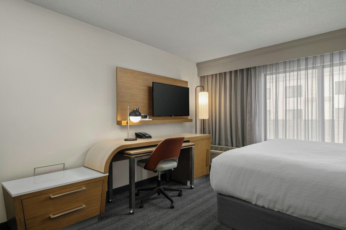 Une chambre d’hôtel comprenant un lit, un bureau avec une chaise, une télévision et une fenêtre avec des rideaux.