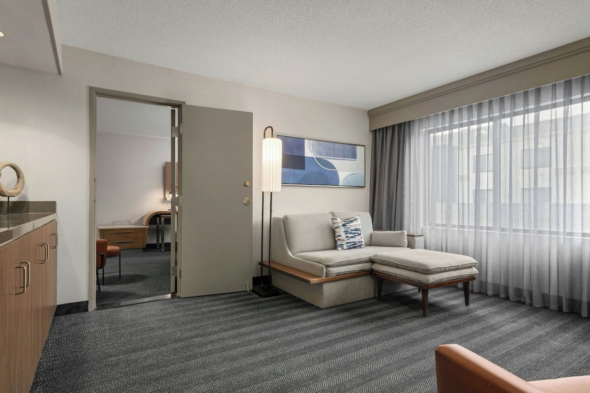 Een hotelkamer met een bank, een bureau, een lamp en een groot raam met gordijnen.