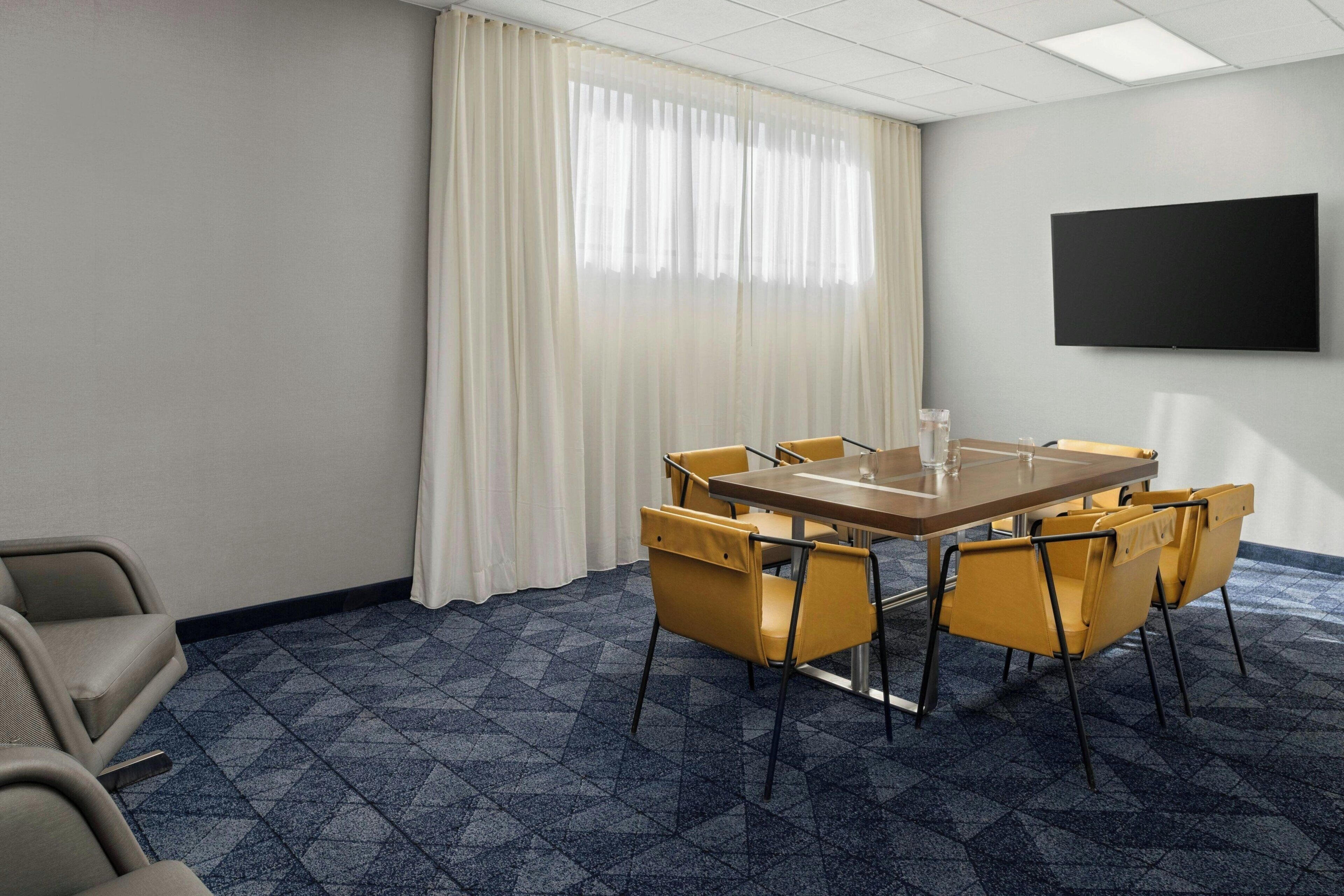 Un salón de conferencias con una gran mesa, sillas amarillas, un televisor de pantalla plana y un ventanal con cortinas blancas.
