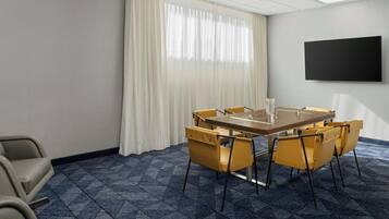 Un salón de conferencias con una gran mesa, sillas amarillas, un televisor de pantalla plana y un ventanal con cortinas blancas.