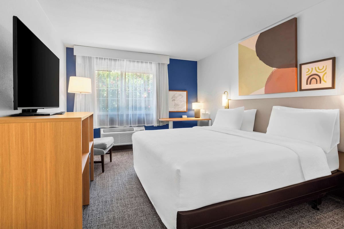 A hotel room with a large bed, a desk with a flat-screen TV, a chair, a desk lamp, and a window with sheer curtains.