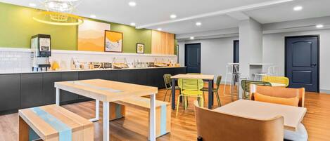 A modern, well-lit café with a wooden floor, a counter with a coffee machine, and seating areas with chairs and tables.