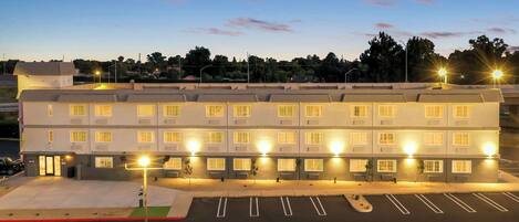 A multi-story hotel with a row of uniformly lit windows, a parking lot with marked spaces, and a flat-roofed structure.