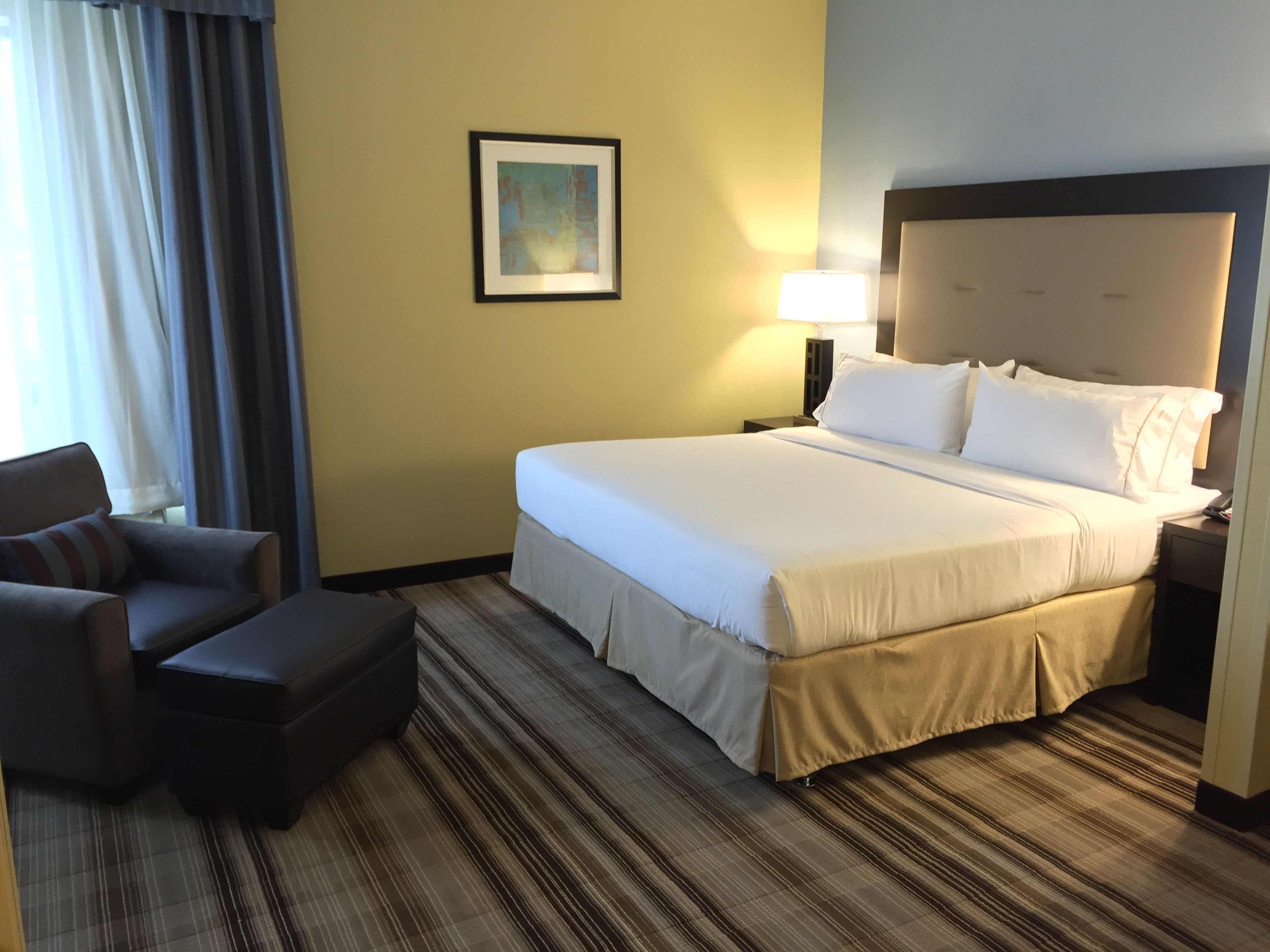 A hotel room with a large bed, a chair, a small table, and a framed picture on the wall.