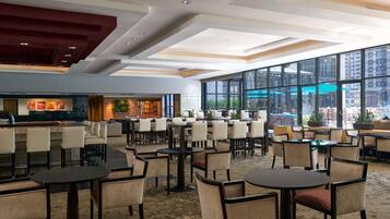 A spacious indoor area with multiple tables and chairs, large windows offering views of the city, and a modern ceiling design.