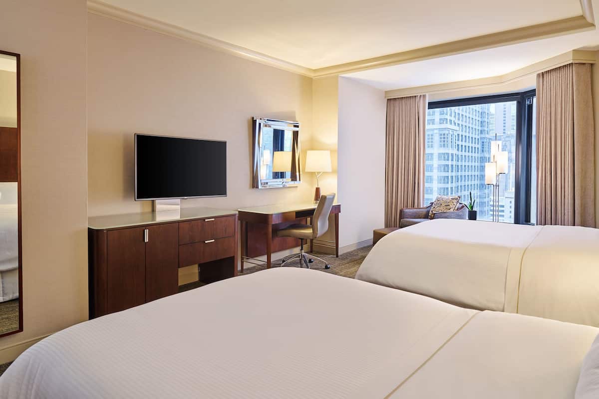 Une chambre d’hôtel avec deux lits, un bureau doté d’un téléviseur à écran plat, une chaise, une lampe et une grande fenêtre avec des rideaux.