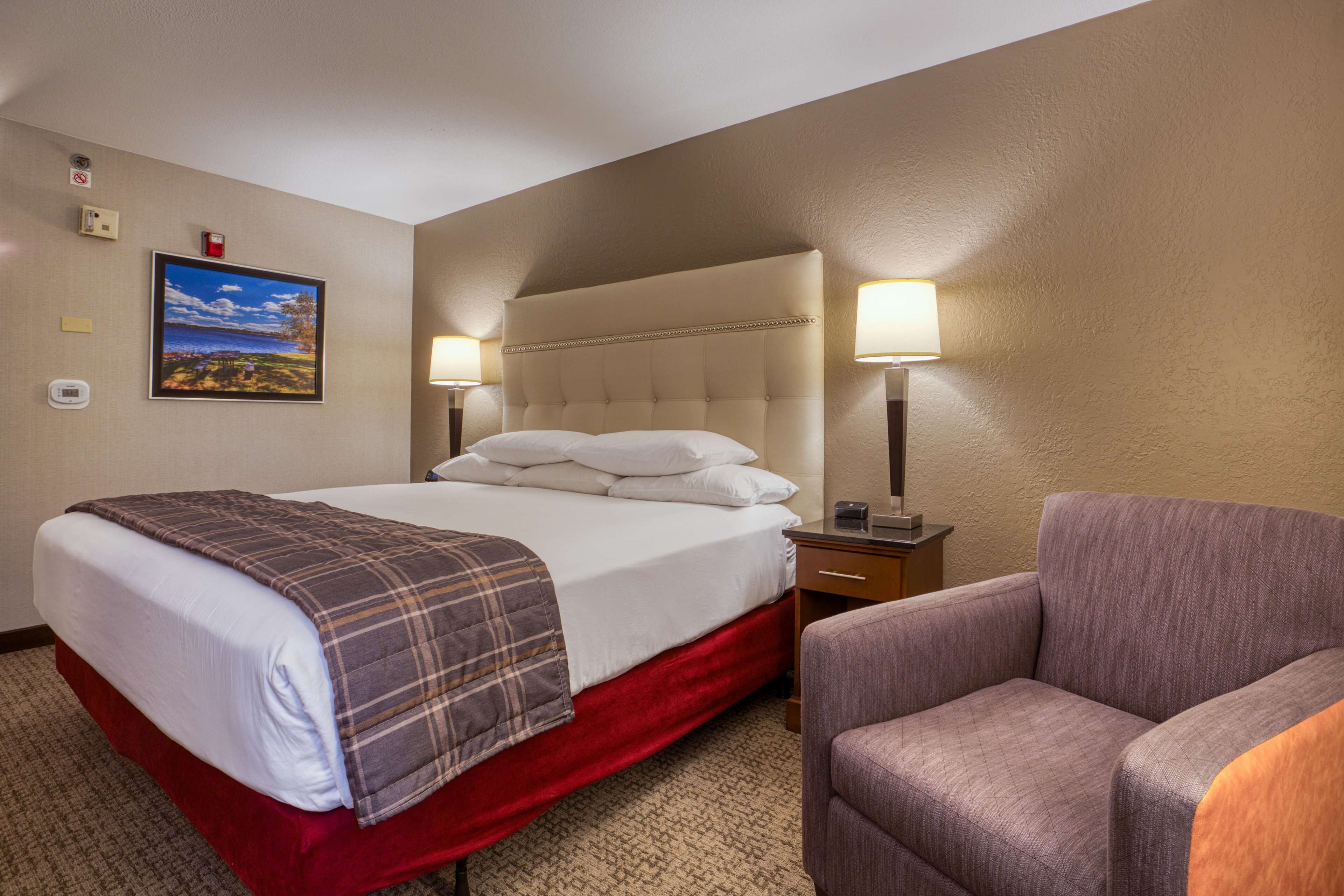 A hotel room with a bed, a chair, a nightstand, and a wall-mounted lamp.