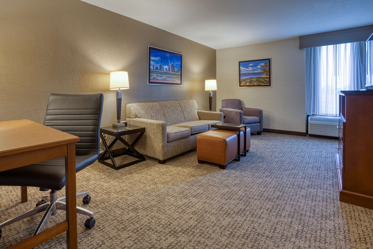A hotel room with a large desk, two chairs, a sofa, and a small table. There are two wall-mounted lamps and two framed pictures on the walls.