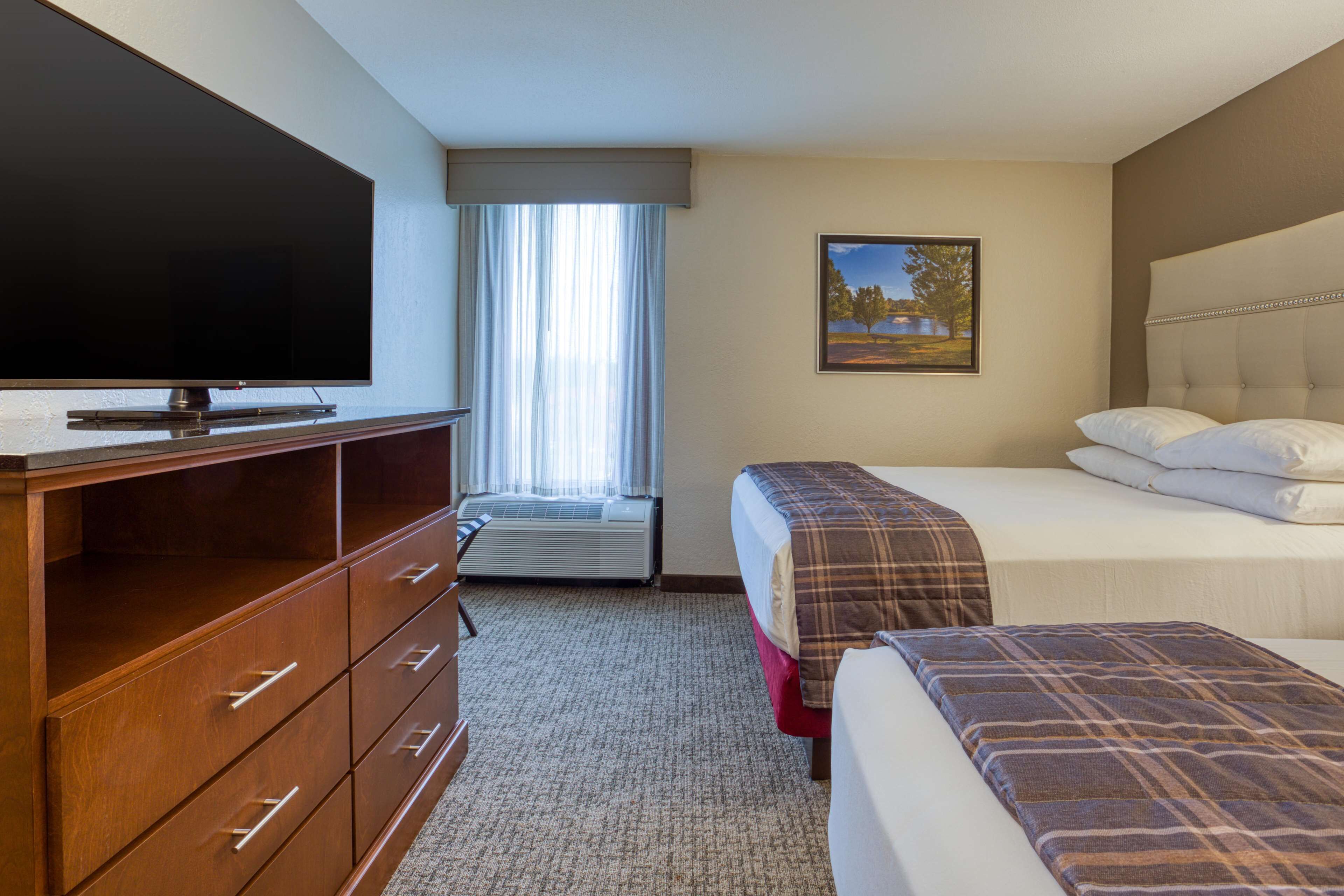 A hotel room with a flat-screen TV, a wooden dresser, a bed with a plaid blanket, a window with curtains, and a framed picture on the wall.