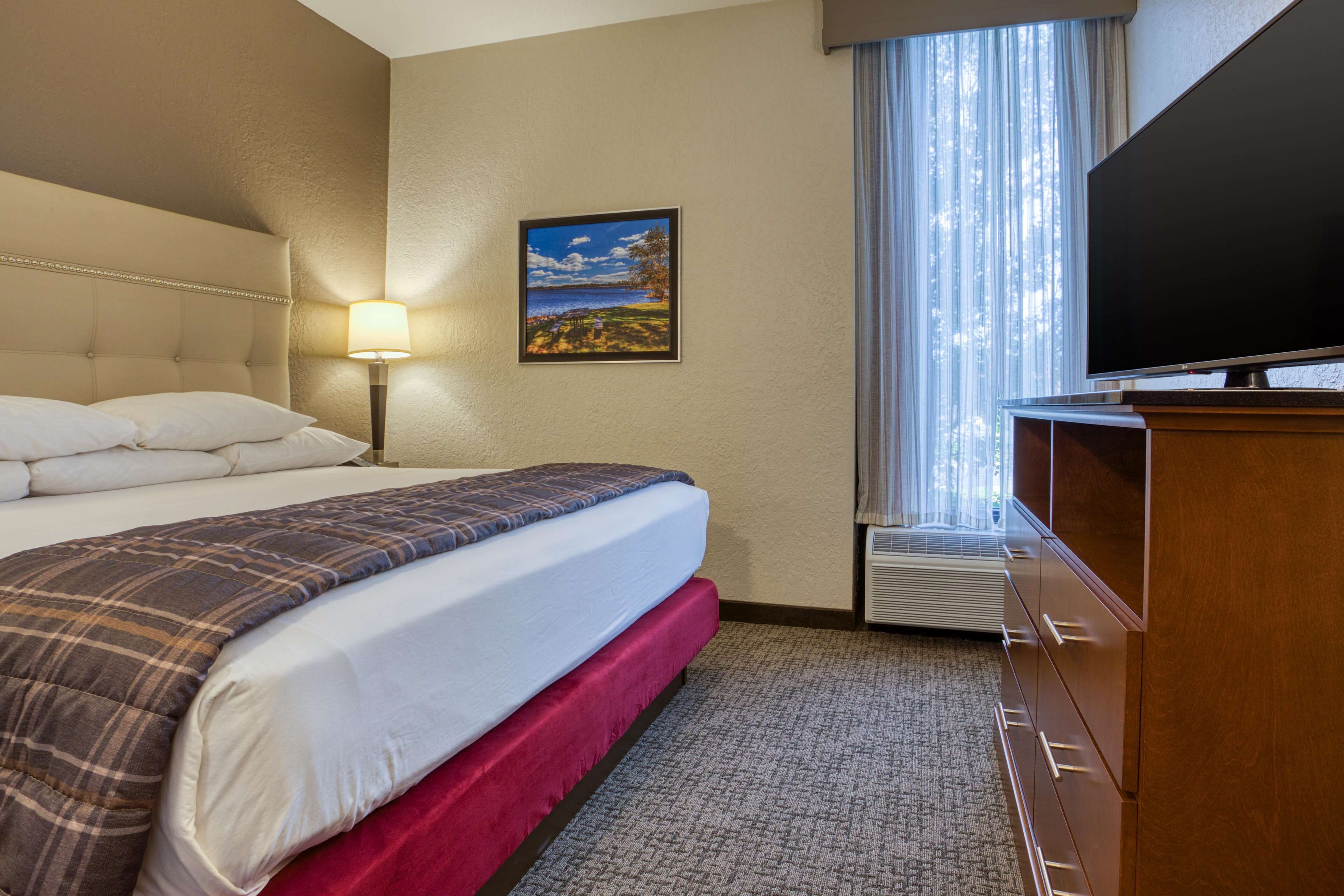 A hotel room with a large bed, a flat-screen TV, a painting on the wall, and a window with curtains.