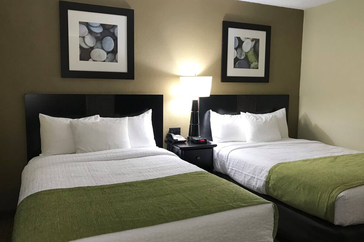 A hotel room with two beds, each with white bedding and a green throw. There are framed artworks on the walls and a nightstand with a phone and a lamp.