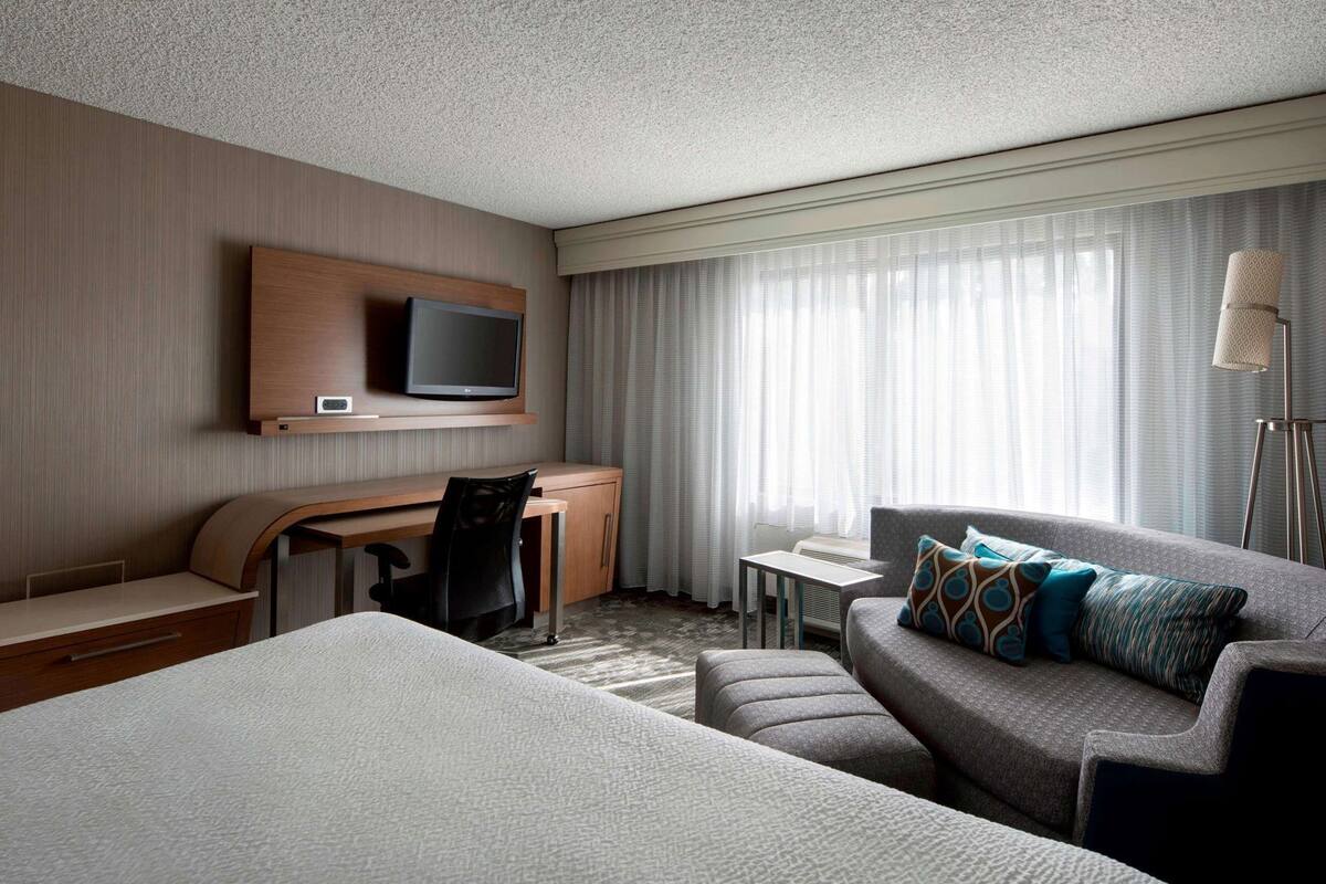 A hotel room with a bed, a desk with a chair, a television, a sofa with pillows, and a floor lamp.