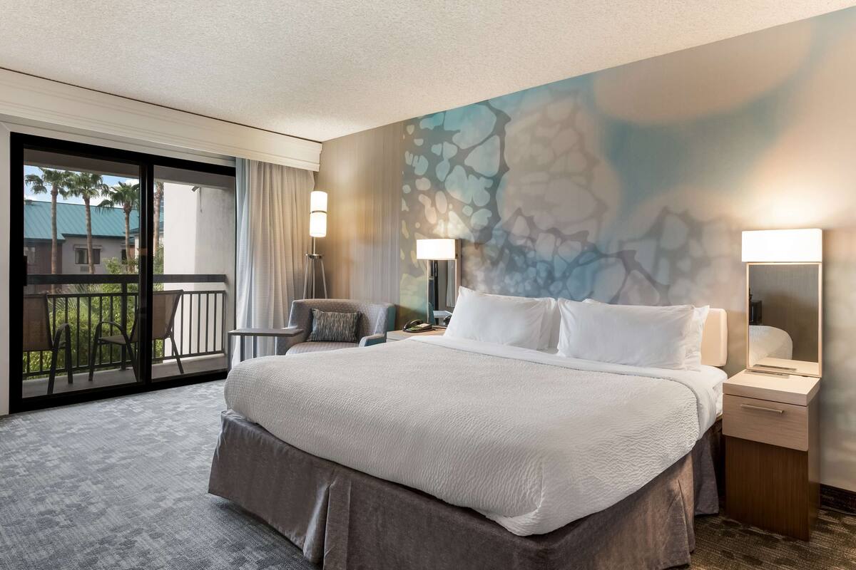 A hotel room with a large bed, a desk with a lamp, a chair, and a balcony with a view of palm trees.