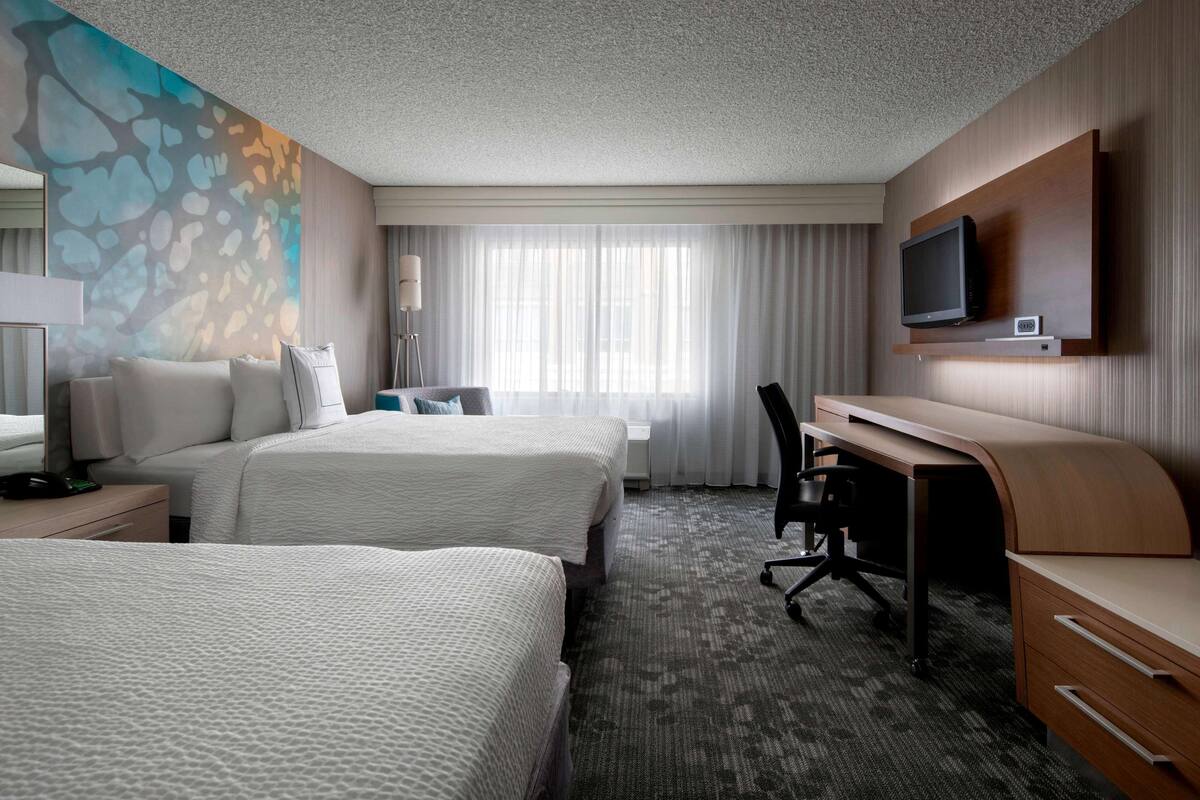 A hotel room with two beds, a desk, a chair, a TV, and a large window with curtains.