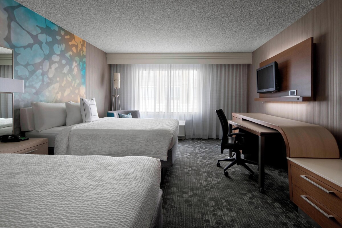 A hotel room with two beds, a desk, a chair, a TV, and a large window with curtains.