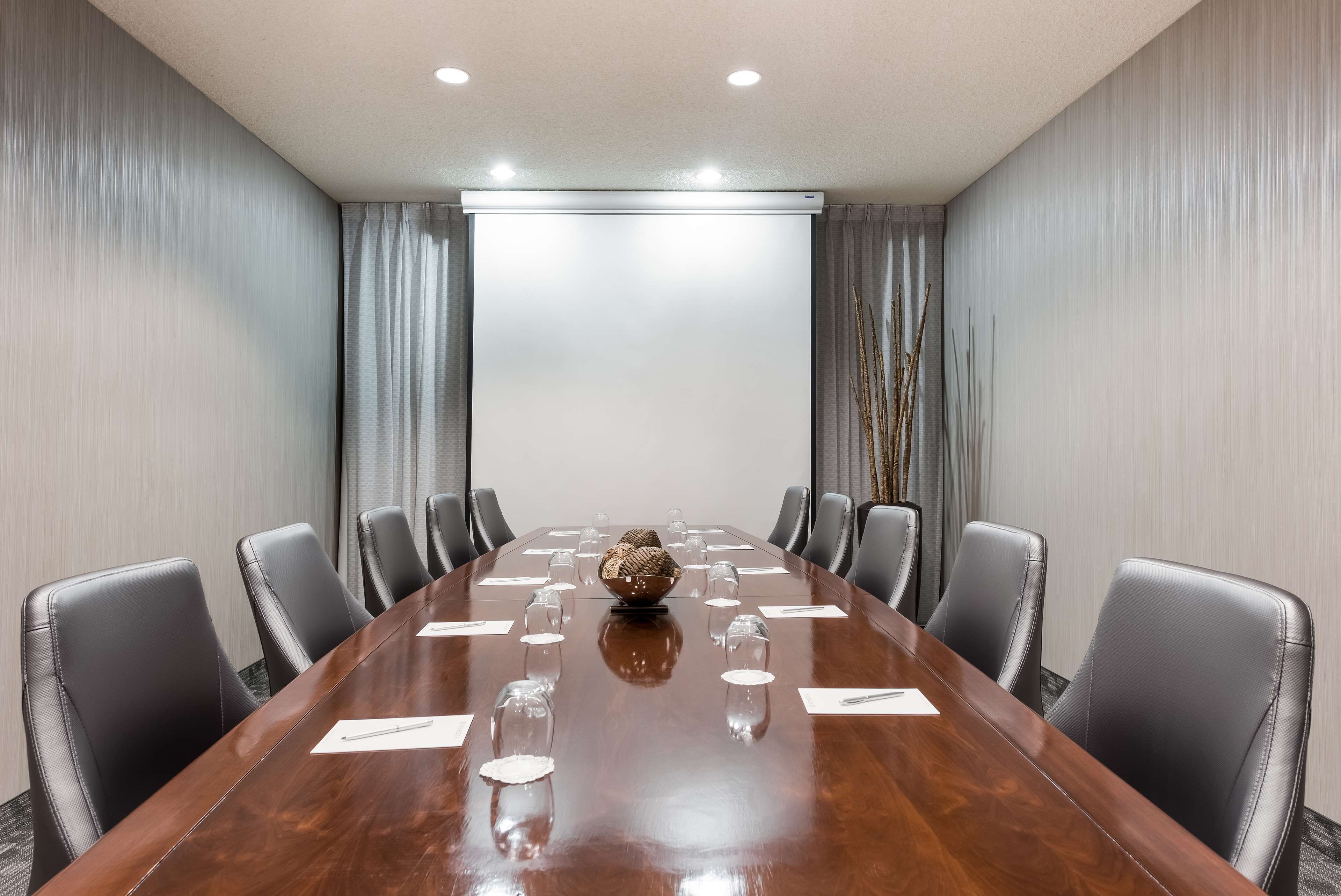 A conference room with a long wooden table, black leather chairs, and a projector screen at the front.