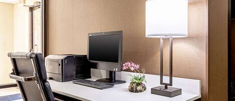 A hotel workspace with a desk, computer, printer, chair, lamp, and a small potted plant.