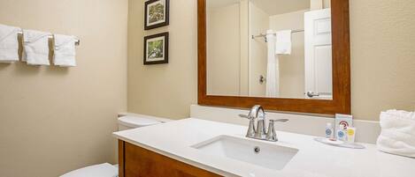 A bathroom with a white sink, a mirror, and a toilet. There are towels hanging on the wall and a framed picture.