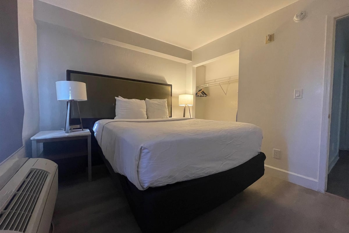 Una habitación de hotel con una cama individual, una mesita de noche con lámpara, un calefactor y un armario con ropa colgada.