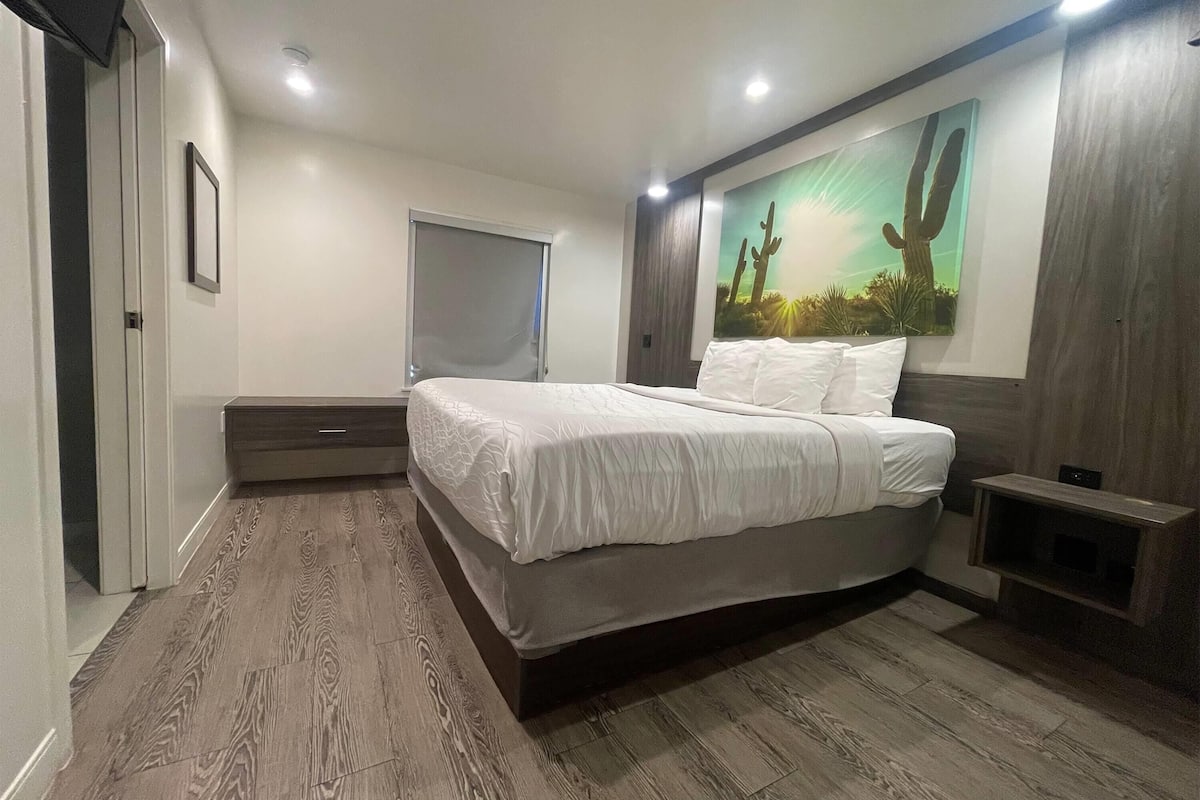 Un dormitorio con cama, mesita de noche, televisión montada en la pared y una pintura de cactus.