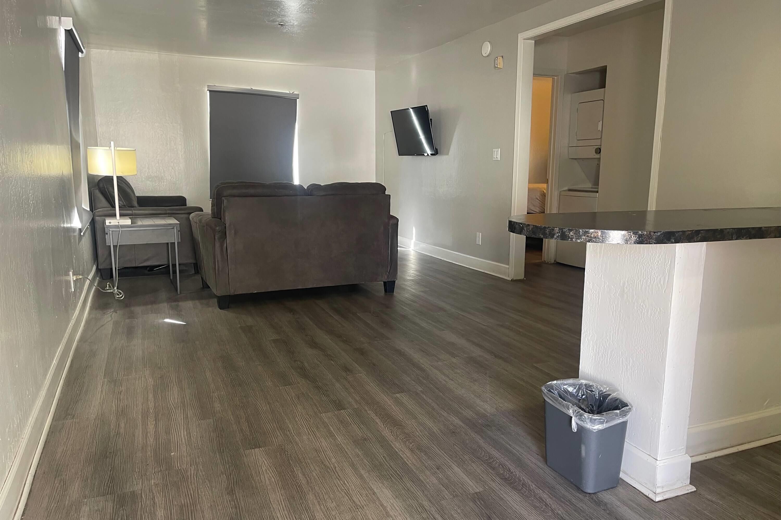 A modern living room with a grey sofa, a small table with a lamp, a flat-screen TV mounted on the wall, and a trash bin.