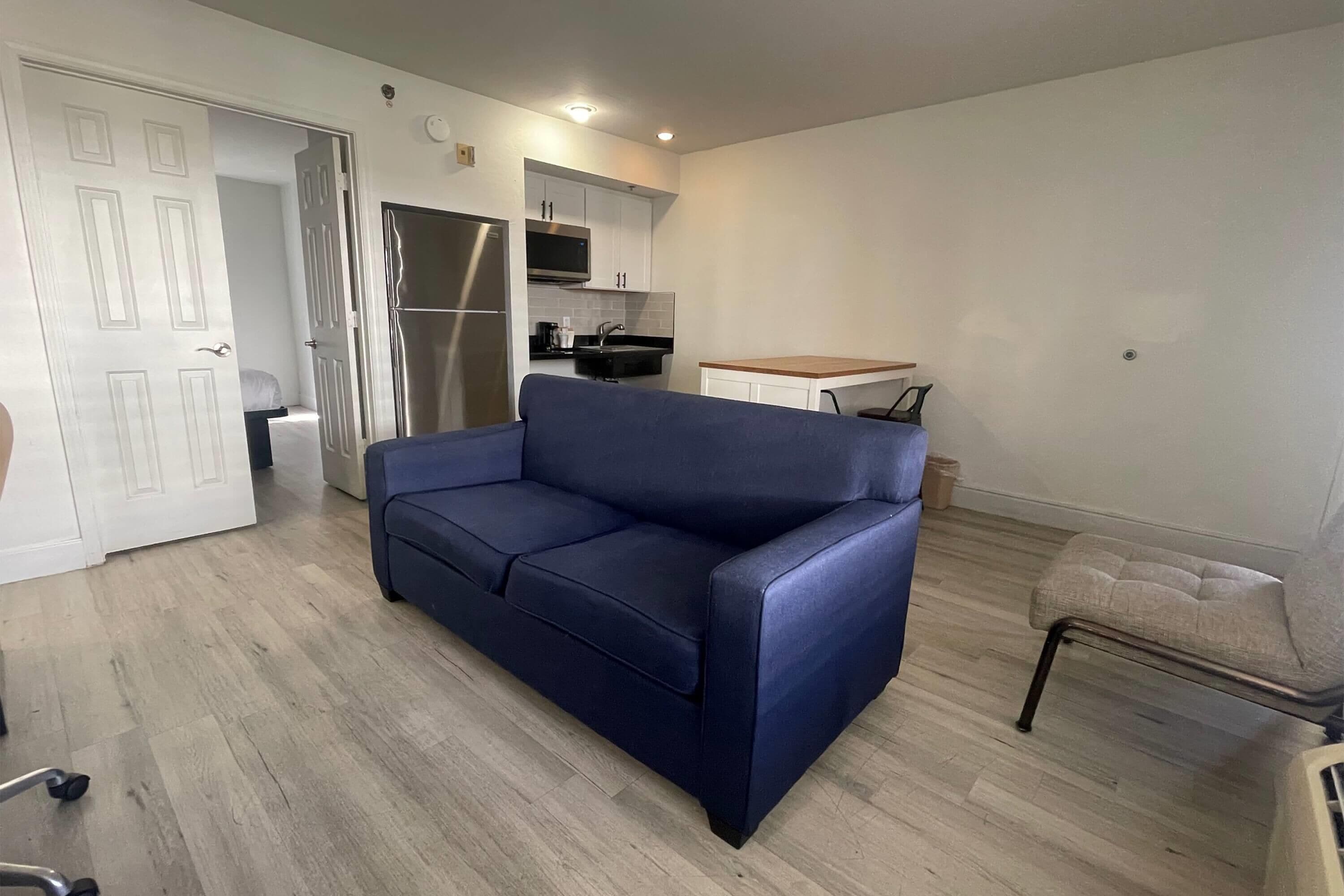 A modern living room with a blue sofa, a wooden coffee table, and a grey armchair. A kitchen with white cabinets and a stainless steel refrigerator is visible in the background.