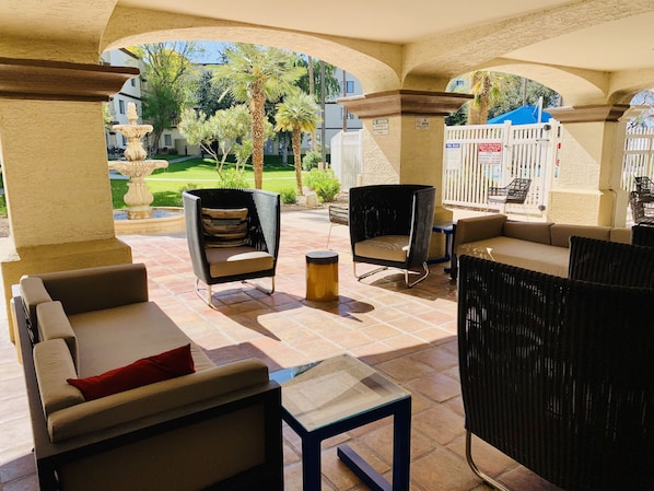 Un patio al aire libre con mobiliario de mimbre, una mesa de vidrio y vistas a una piscina y follaje.
