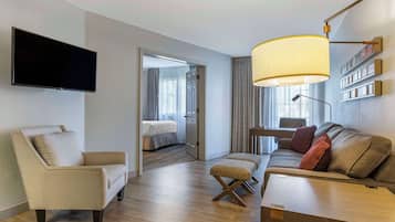 A hotel room with a sofa, armchair, and ottoman, a flat-screen TV mounted on the wall, and a large window with curtains.