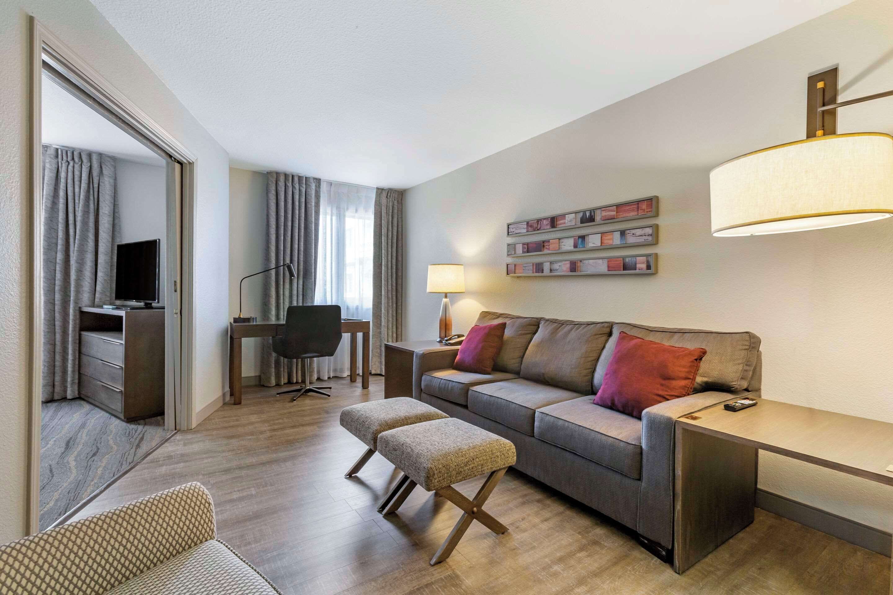 Ein modernes Wohnzimmer mit einer Couch, einem Hocker und einem Schreibtisch mit Stuhl. In der Ecke befindet sich ein Fernseher, eine Lampe und eine an der Wand befestigte Ablage mit Bildern.