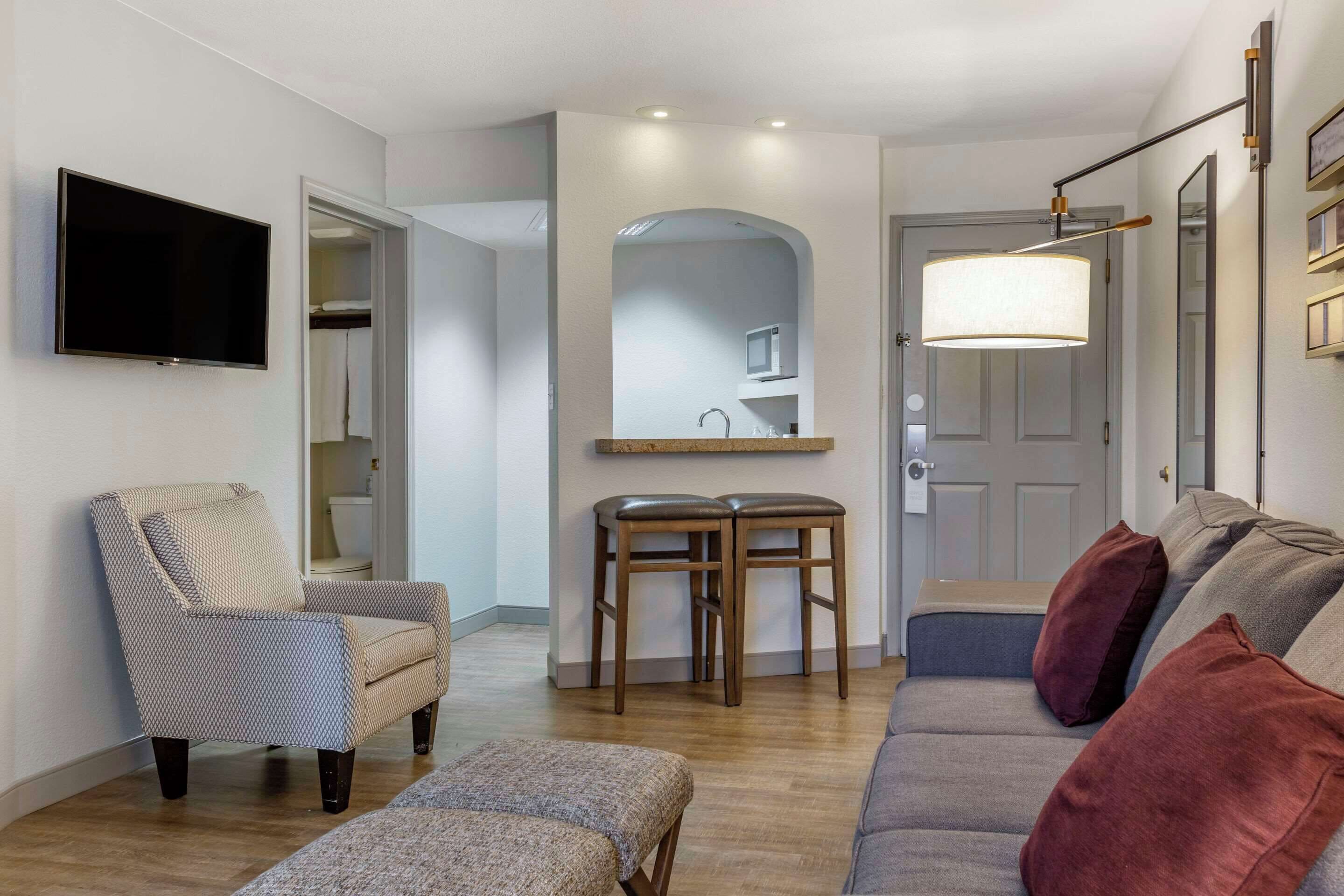 Una sala de estar con sofá, sillón, reposapiés, televisión y un comedor con taburetes y mesa.