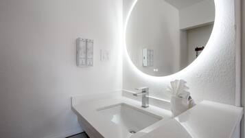 Une salle de bain moderne avec un lavabo blanc, un miroir doté d’un éclairage circulaire et un distributeur de serviettes.