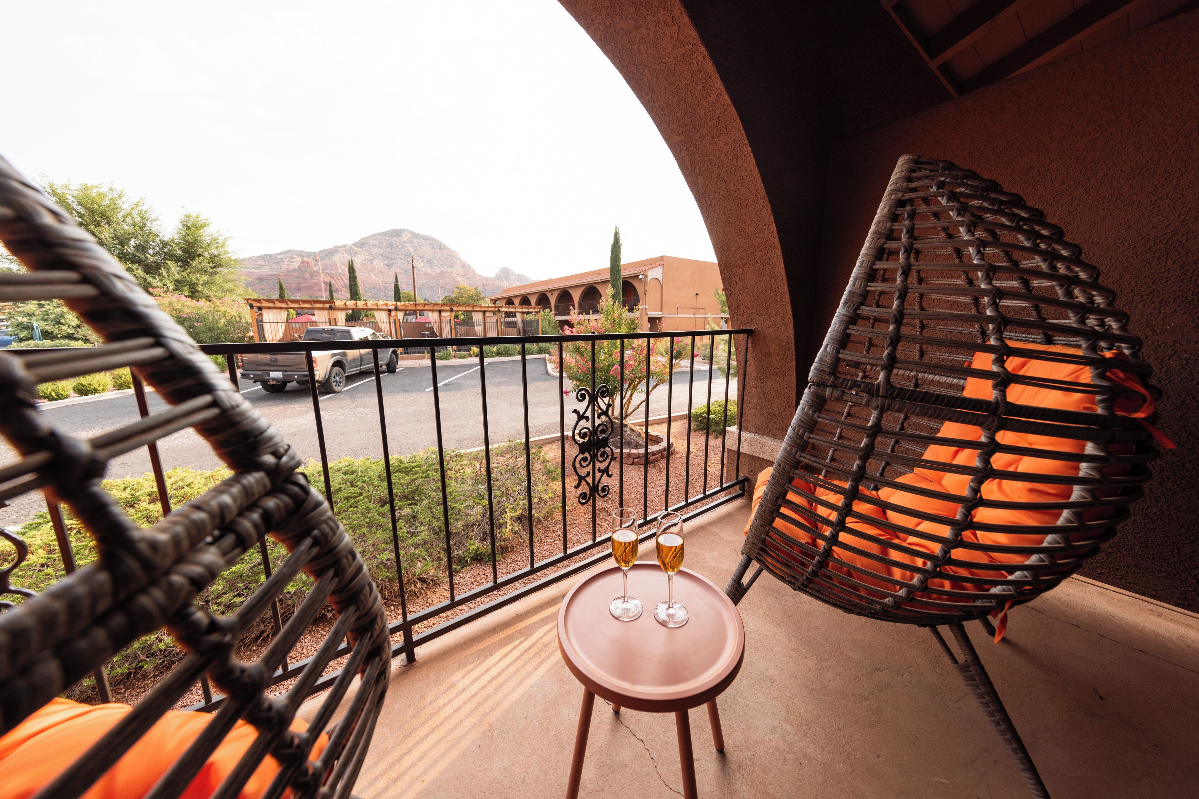 A balcony with a wicker chair, a small table with two glasses, and a view of a parking lot and mountains.