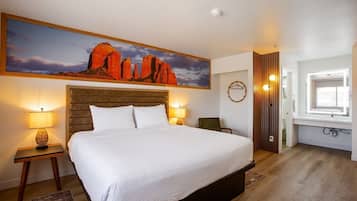 Un dormitorio con una cama grande, dos lámparas de noche, un cuadro en la pared que representa un paisaje desértico, un cabecero de madera, una pequeña silla y una ventana con vistas a la habitación.