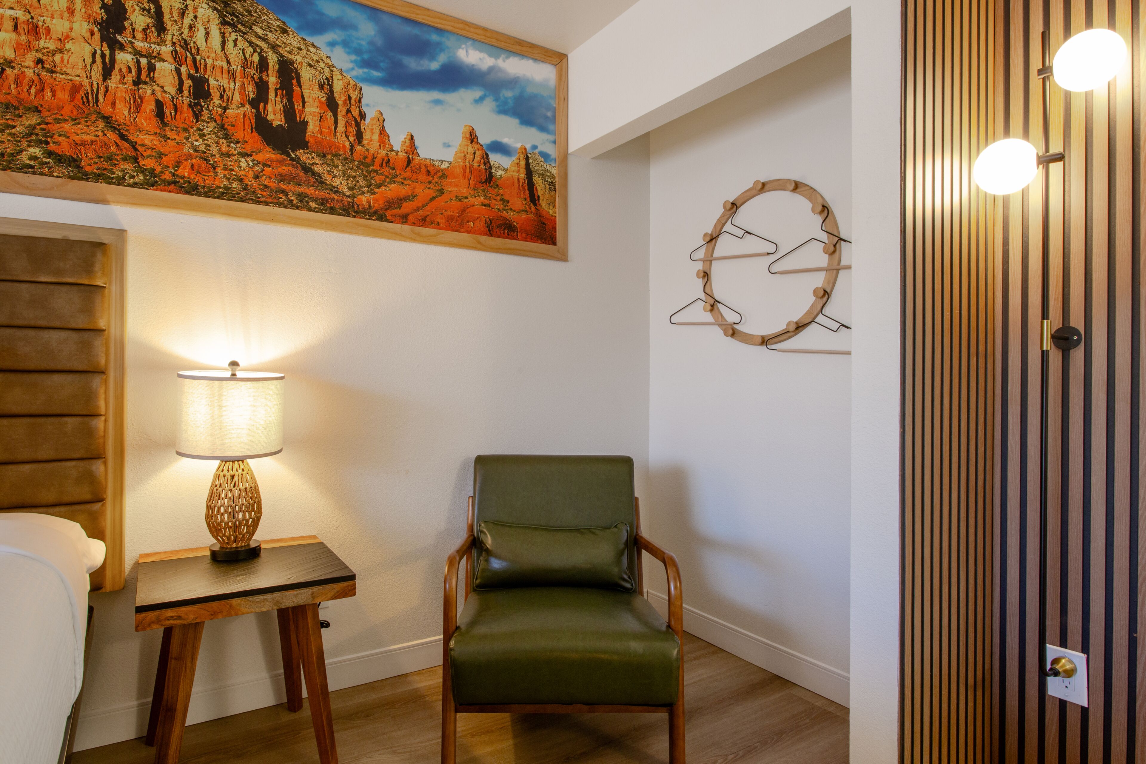 Ein Zimmer mit Holzboden, einem grünen Stuhl, einem Tisch mit Lampe, einem Gemälde einer Wüstenlandschaft und einer Wandleuchte.