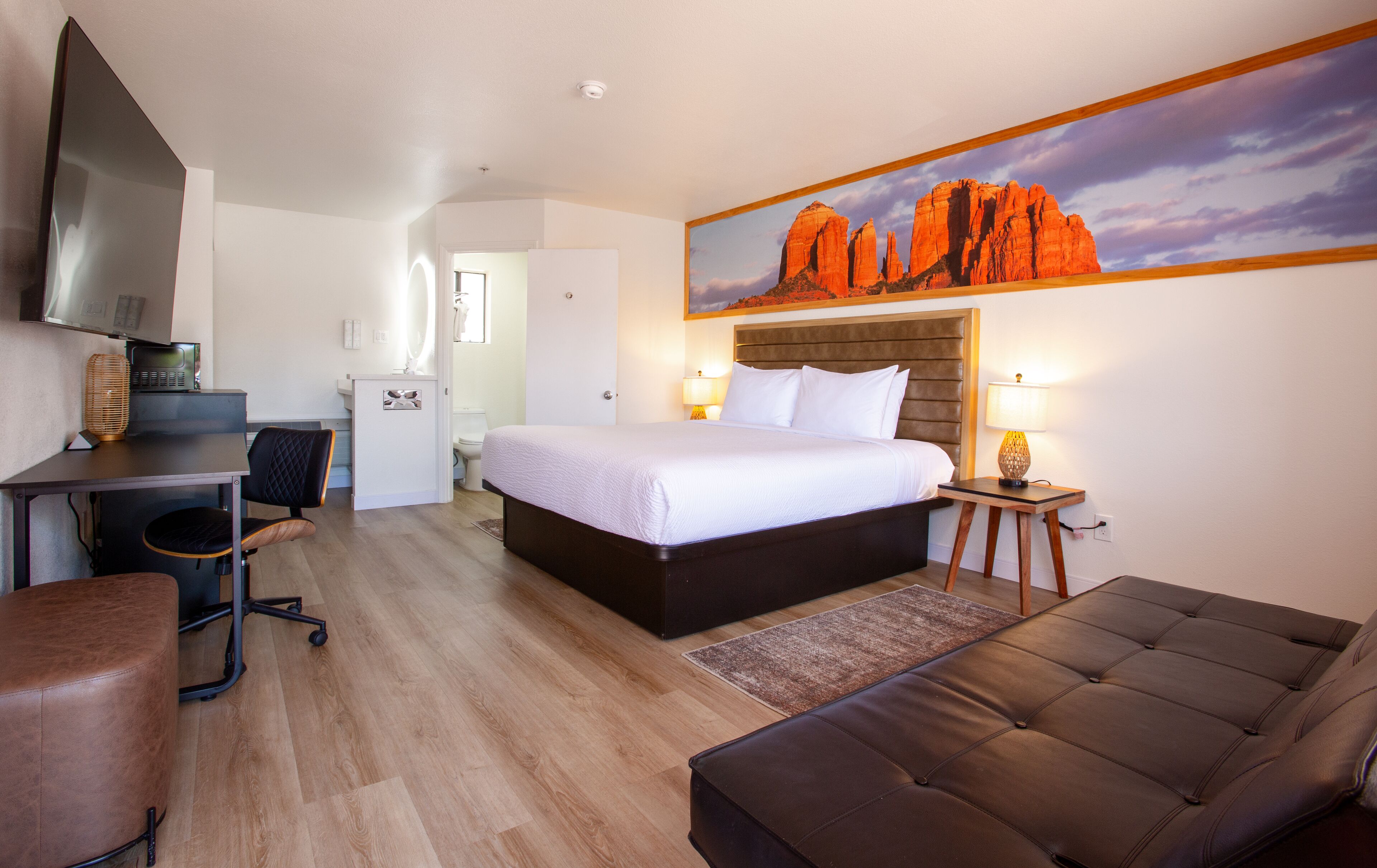 A hotel room with a bed, a desk, a chair, a sofa, a painting of red rock formations, and a television.