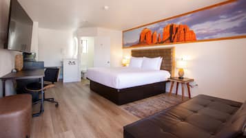 A hotel room with a bed, a desk, a chair, a sofa, a painting of red rock formations, and a television.